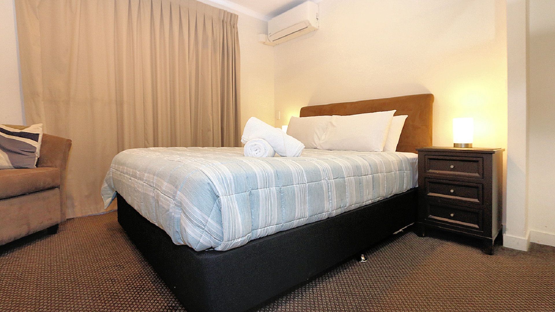 A hotel room with a bed, a nightstand with a lamp, a chair, and a window with curtains.
