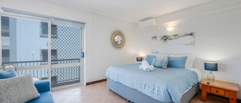 Un dormitorio con cama, mesita de noche, lámpara, espejo en la pared y balcón con vistas a edificios.