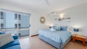 A bedroom with a bed, a nightstand, a lamp, a wall-mounted mirror, and a balcony with a view of buildings. - Shoreline Apartments - Unit 10 (Bargara)