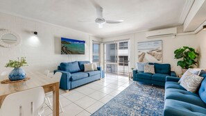 A spacious living area with a ceiling fan, two blue sofas, a dining table, and a large window with a view. - Shoreline Apartments - Unit 10 (Bargara)
