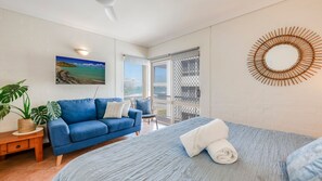 A room with a blue sofa, a bed with a blue bedspread, a large window, a wall-mounted circular wooden decoration, and a painting of a beach. - Shoreline Apartments - Unit 10 (Bargara)