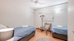 A bedroom with two beds, a ceiling fan, a floor lamp, and a small table with a chair. - Shoreline Apartments - Unit 10 (Bargara)