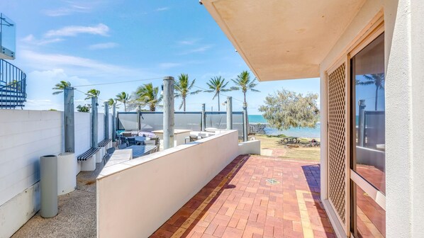 A covered outdoor patio with a view of the beach, outdoor seating, and palm trees. - Shoreline Apartments - Unit 10 (Bargara)
