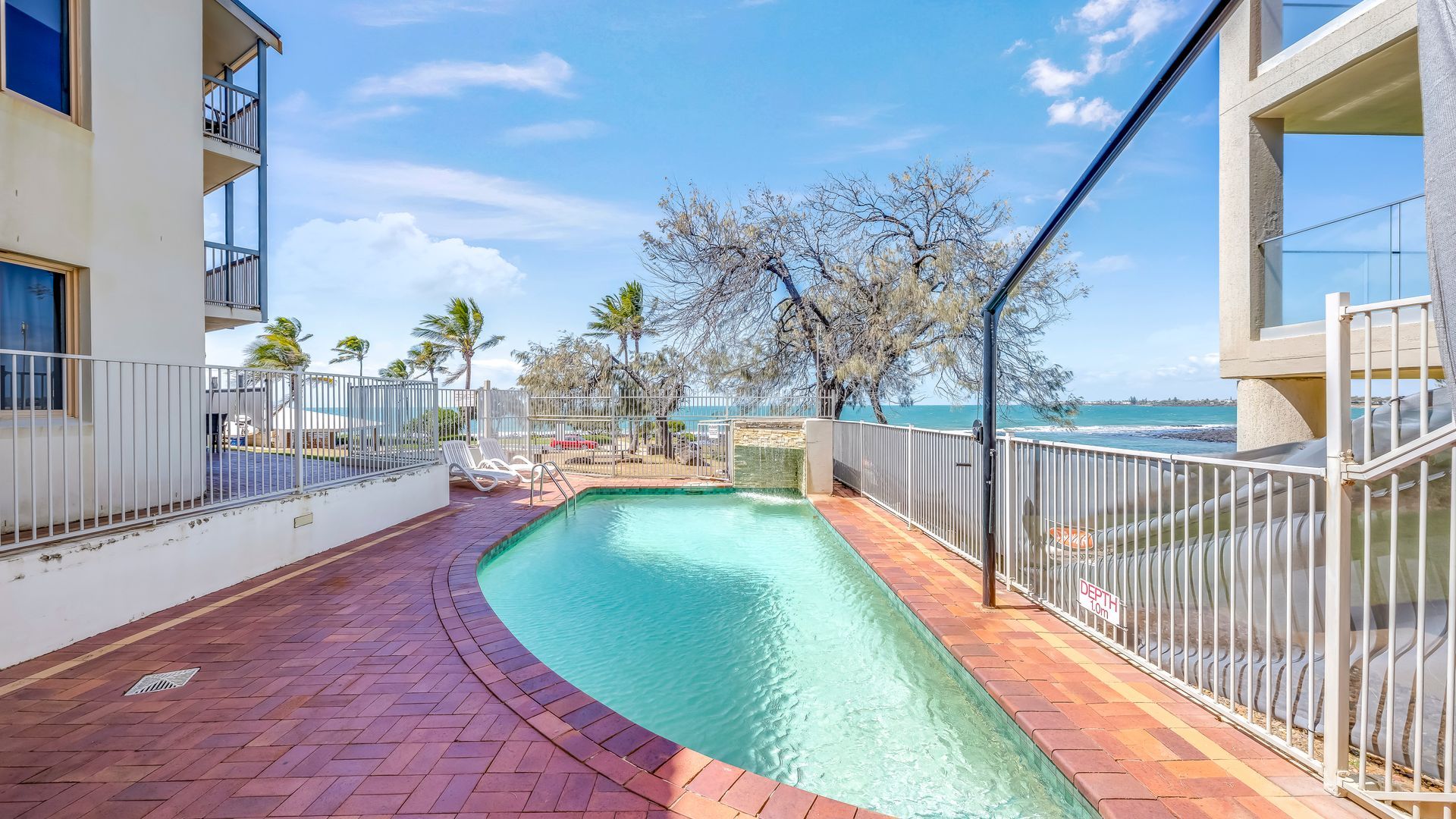 A swimming pool with a clear view of the ocean, surrounded by a white fence, and a brick pathway leading to the pool.