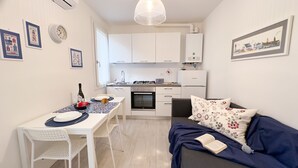 A compact kitchen with white cabinets, a gas stove, and a built-in oven. There is a dining table with chairs, a sofa with a floral cushion, and a framed picture on the wall. - Maison De La Brodeuse by Ca-sa Check (Chioggia)