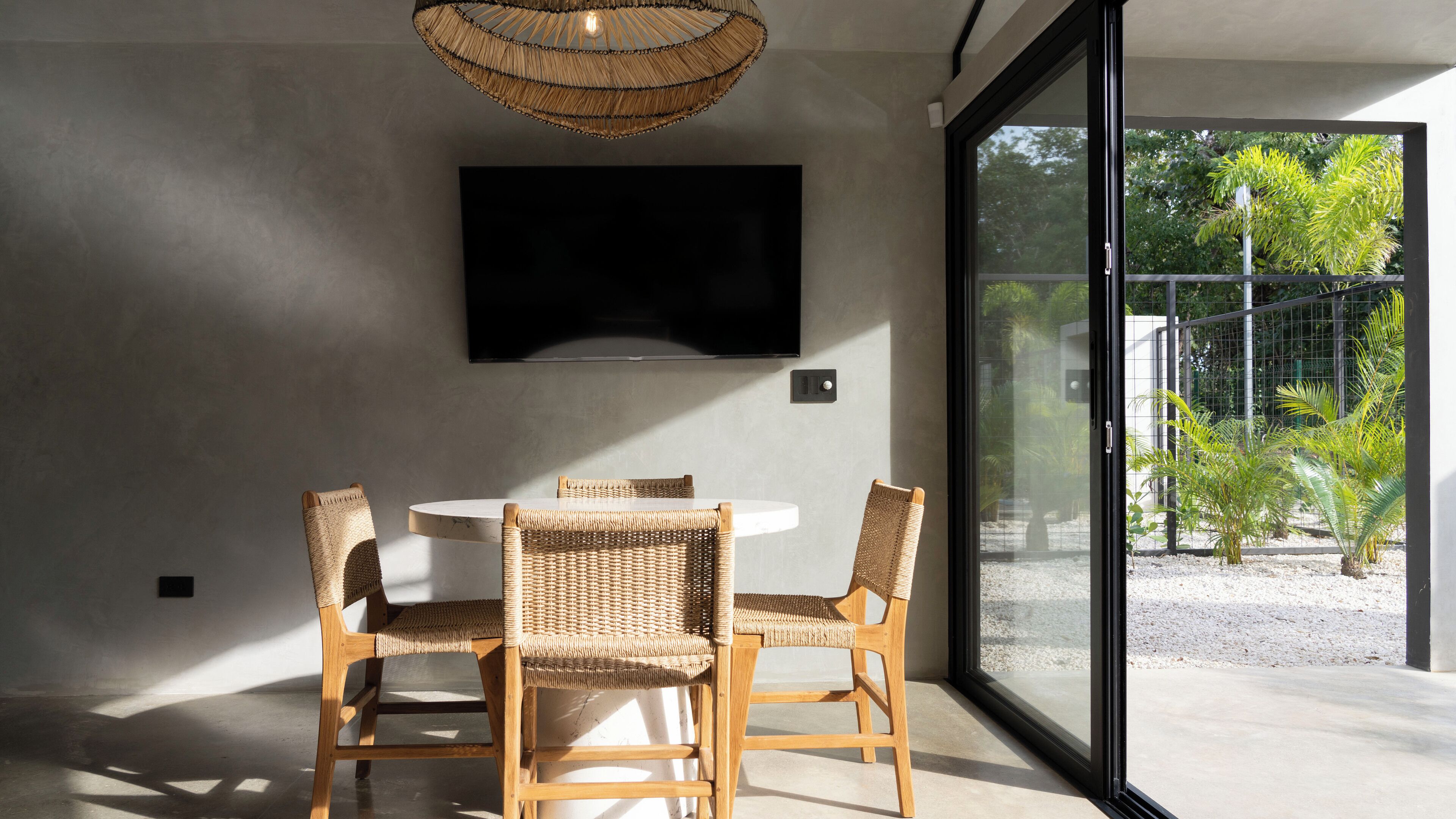 A modern interior with a round table and four wicker chairs, a mounted TV, and a large glass door leading to an outdoor area with greenery.