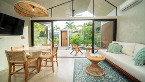 A modern living room with a sofa, a dining table with chairs, a TV mounted on the wall, and a large window offering a view of the outdoors. - Selvatico Tamarindo Villas (Tamarindo)