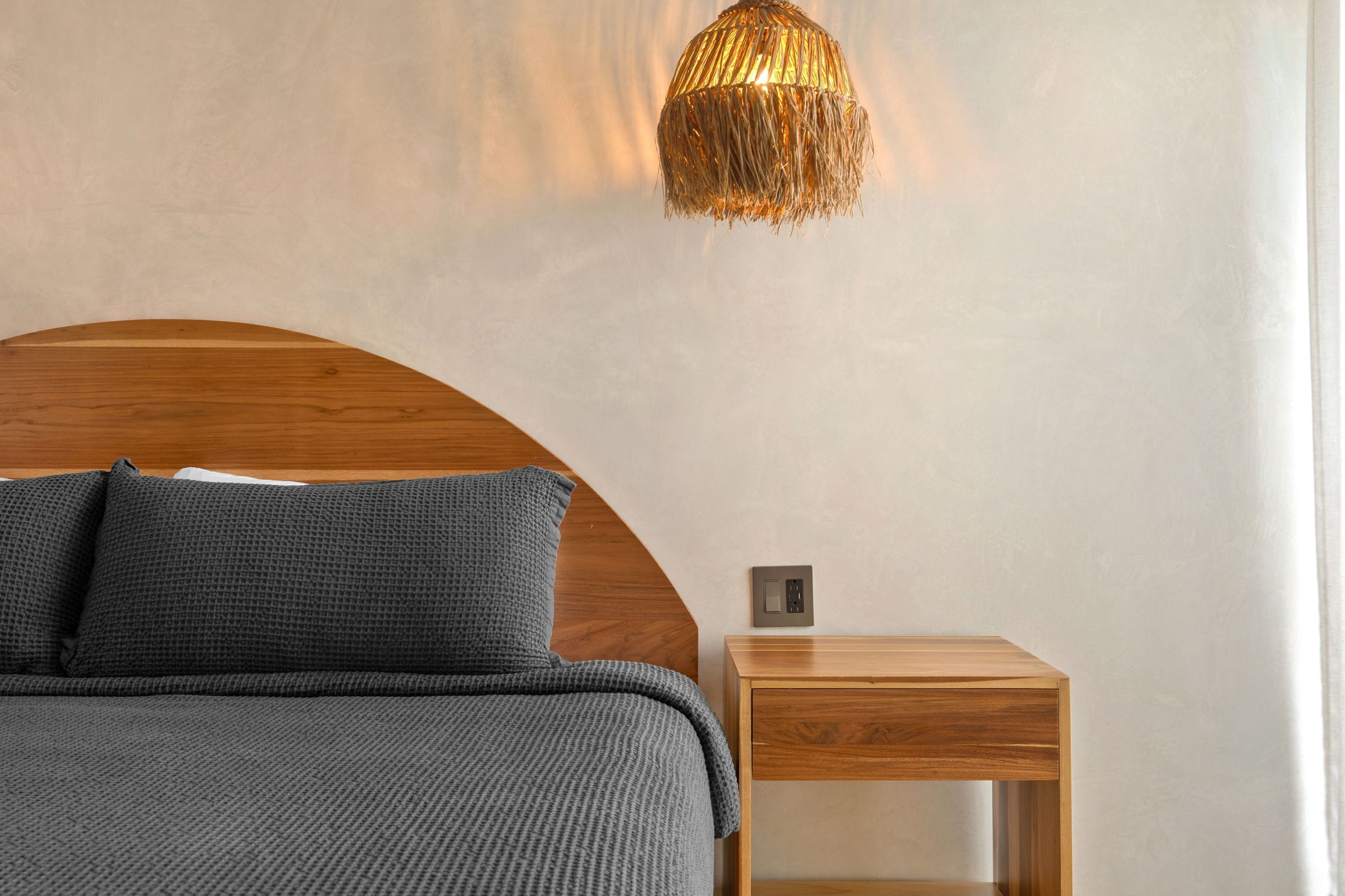 A bedroom with a wooden headboard, a bed with grey bedding, a bedside table, and a wall-mounted light fixture.