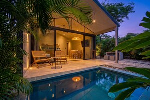 A modern house with a swimming pool, outdoor seating, and a kitchen visible through glass doors.