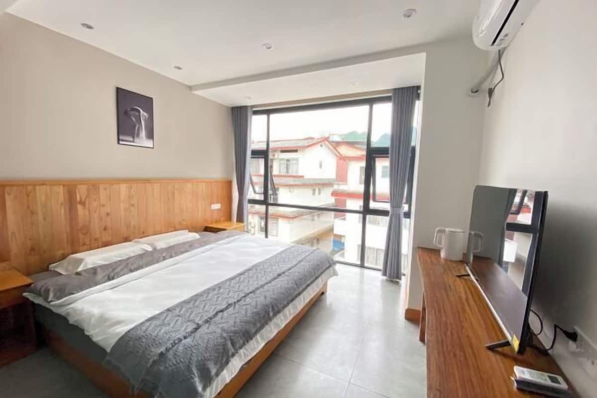 A bedroom with a bed, a wooden headboard, a TV, and a large window with a view of buildings.