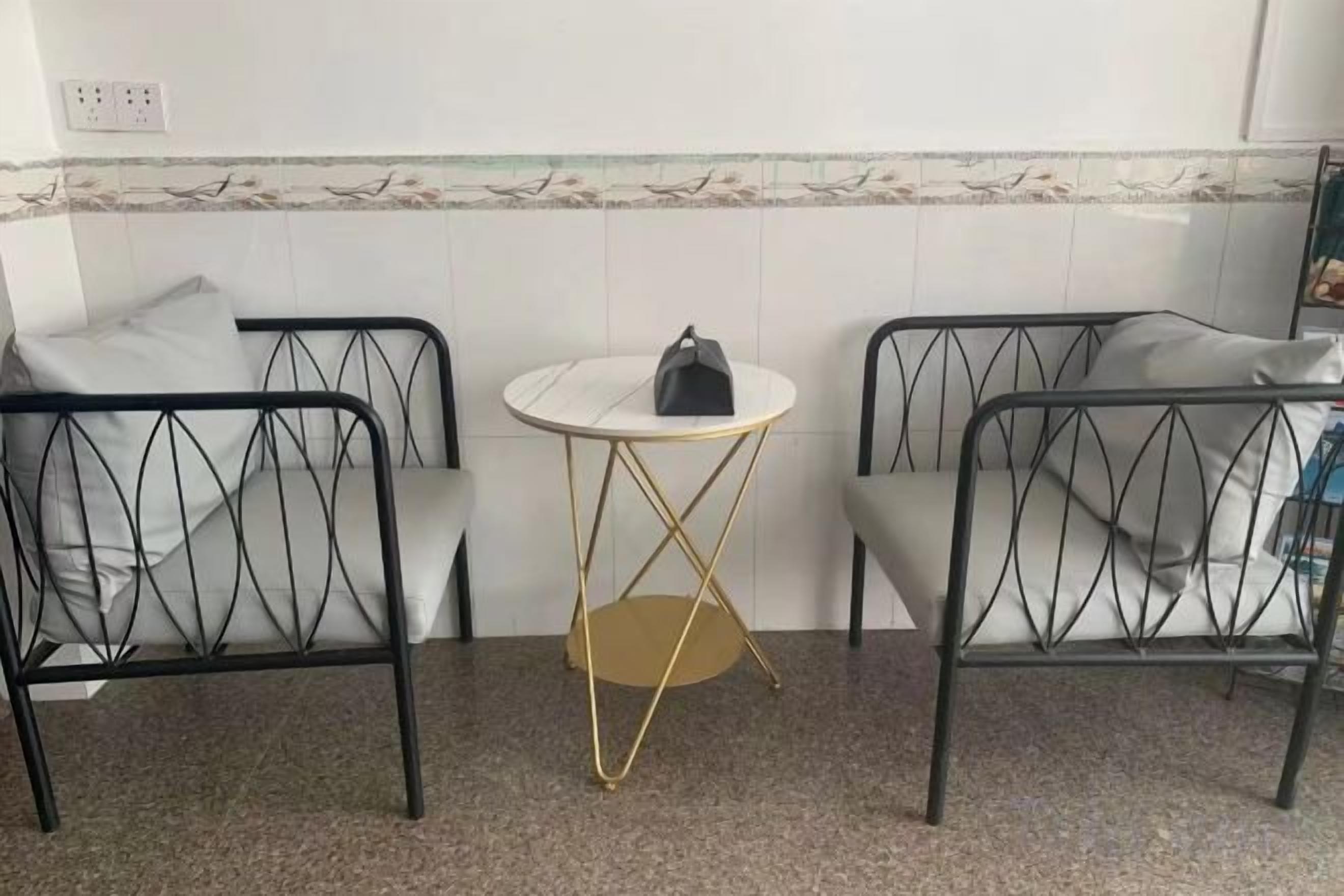 Habitació amb dues cadires de metall negre, una taula rodona petita amb base daurada i una paret blanca amb un adorn decoratiu.