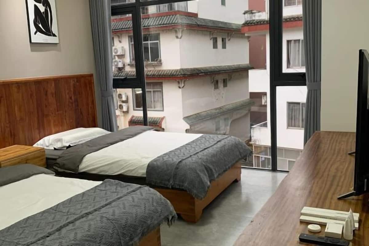 A hotel room with two beds, a wooden headboard, a large window with a view of buildings, and a desk with a television.