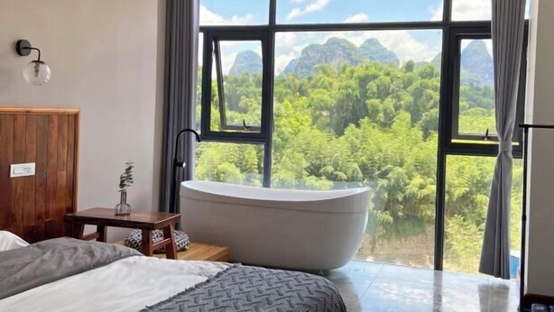 A bedroom with a large window, a bathtub, a wooden cabinet, and a small table with a plant. - Qiqi Xiaozhan (Yangshuo Impression Liusanjie Scenic Area Shop) (Yangshuo)