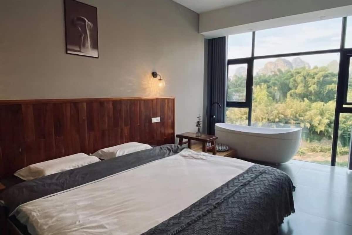 A modern bedroom with a large bed, a wooden headboard, a wall-mounted light, and a view of greenery through large windows.