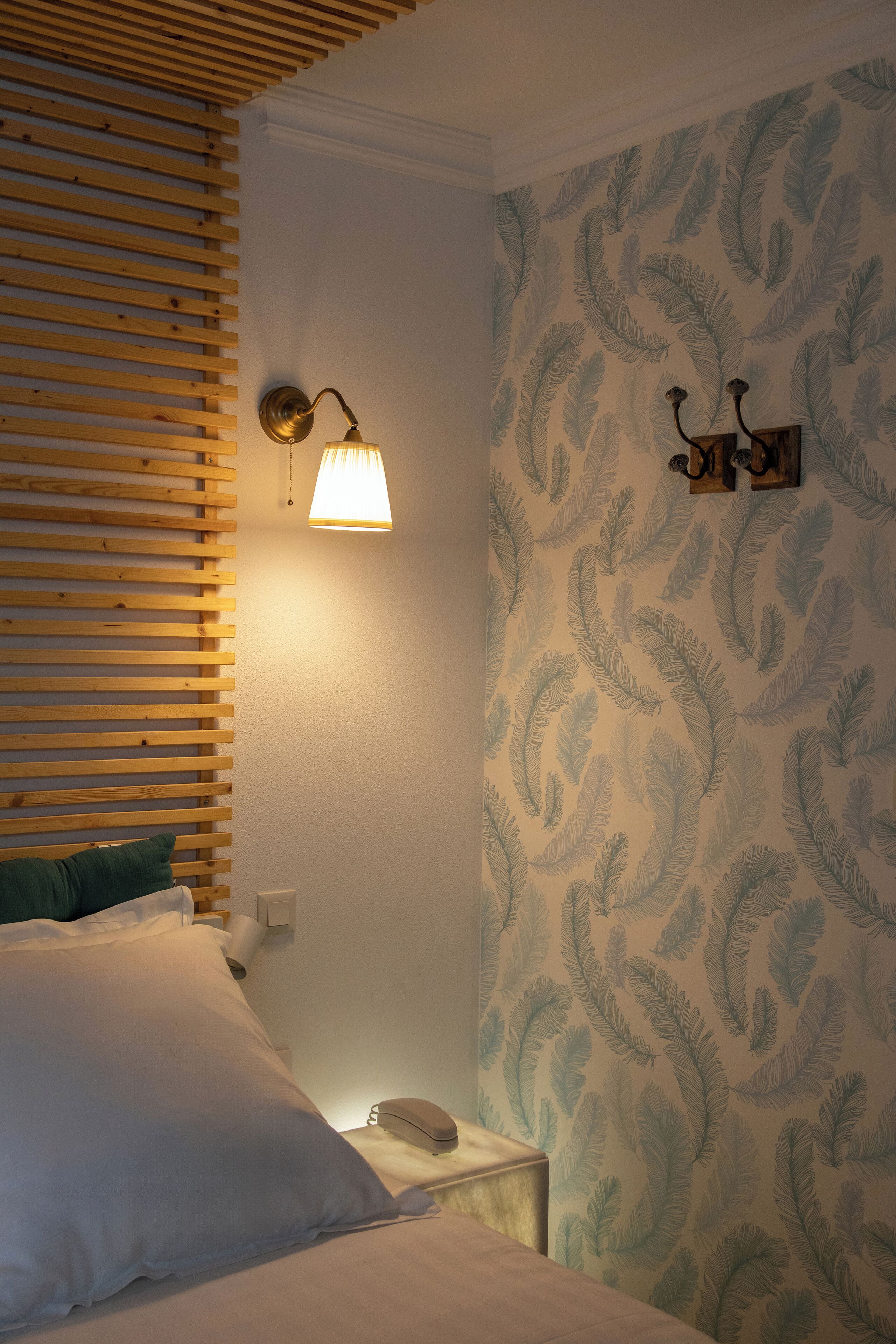 A bedroom with a wooden slat headboard, a bed with white bedding, a nightstand with a lamp, and a wall with a leaf-patterned wallpaper.