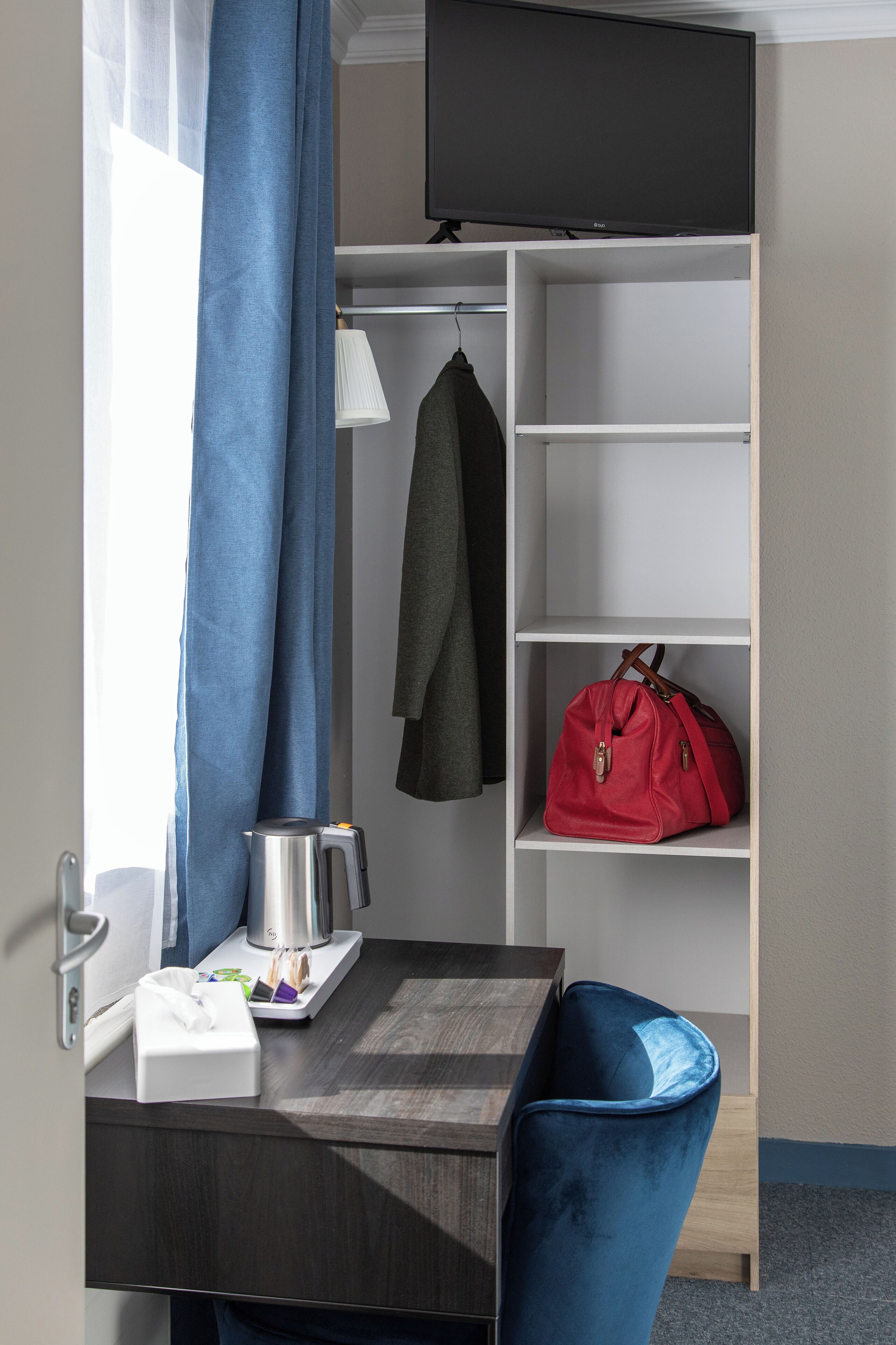 A hotel room with a desk, a chair, a TV mounted on the wall, a shelf with a red bag, and a coat hanging on the wall.