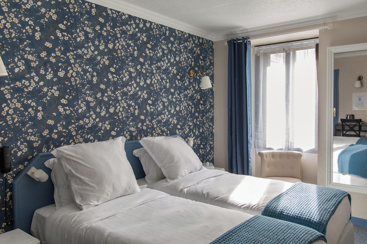 A hotel room with two beds, a floral wallpaper, a window with curtains, a bedside lamp, and a view of another room.