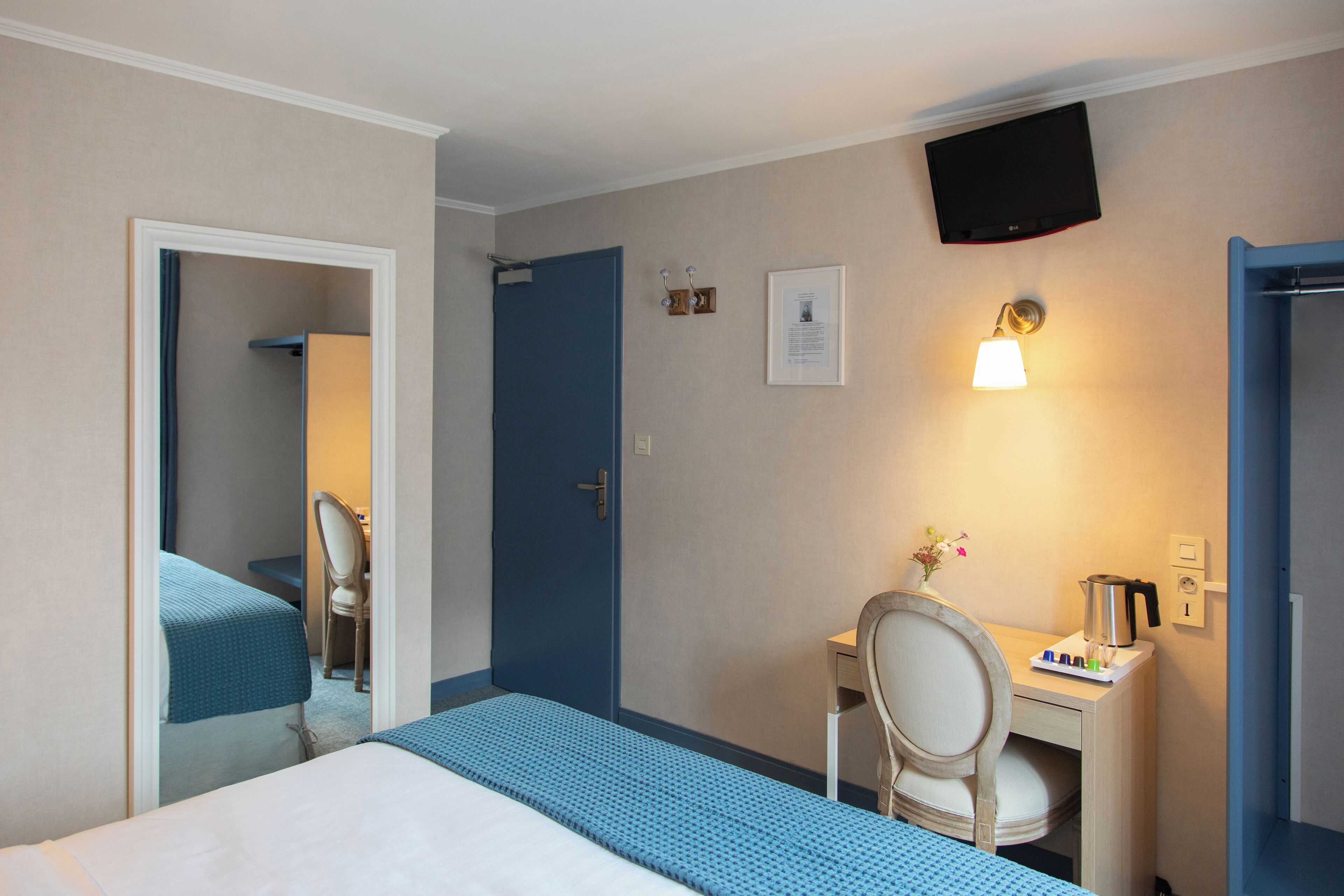 Una habitación de hotel con cama, escritorio y silla, televisor montado en la pared y una puerta.