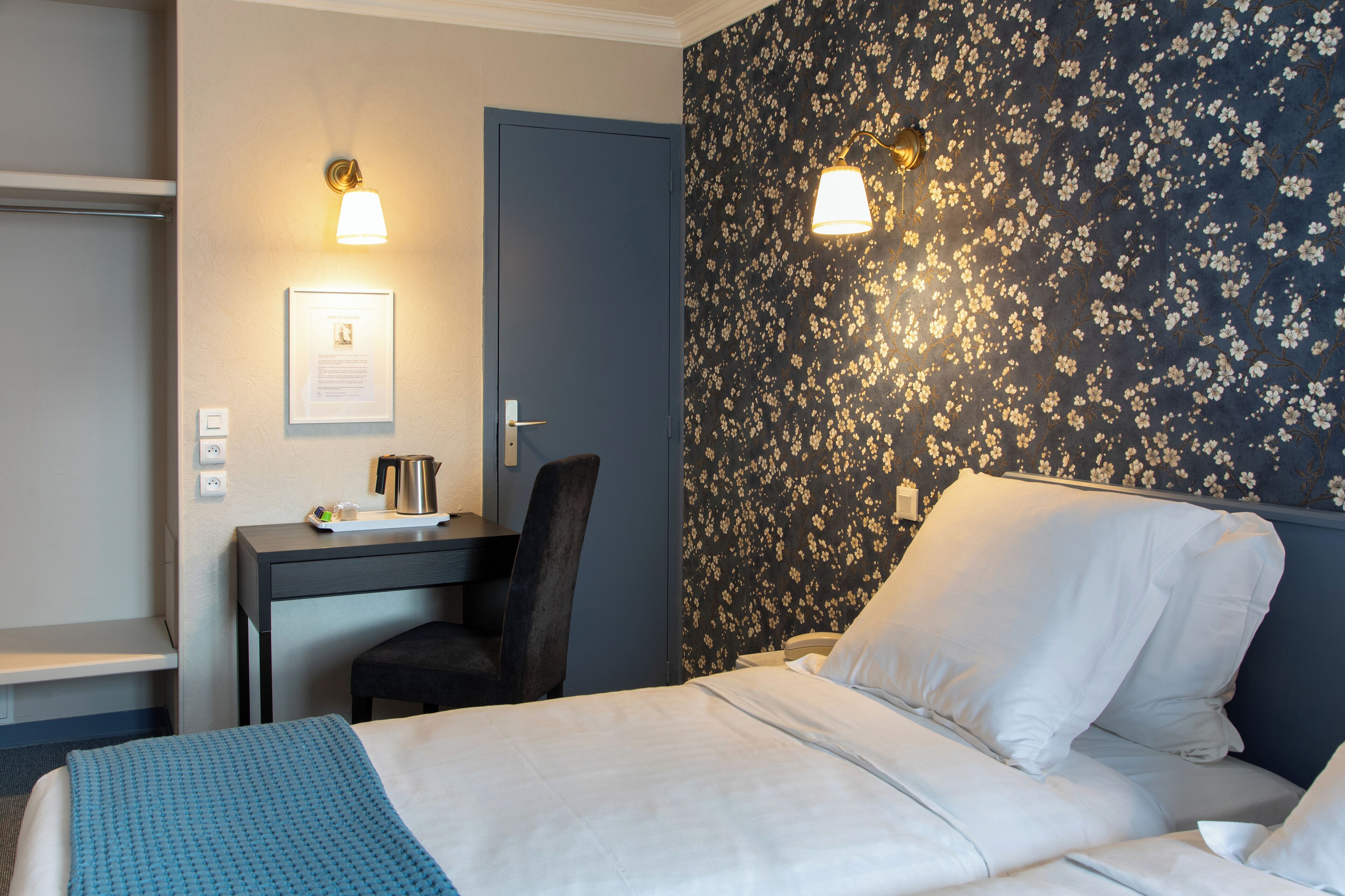 Habitación de hotel con cama, escritorio y silla. Se observa un cuadro enmarcado en la pared y una lámpara fijada a la pared.