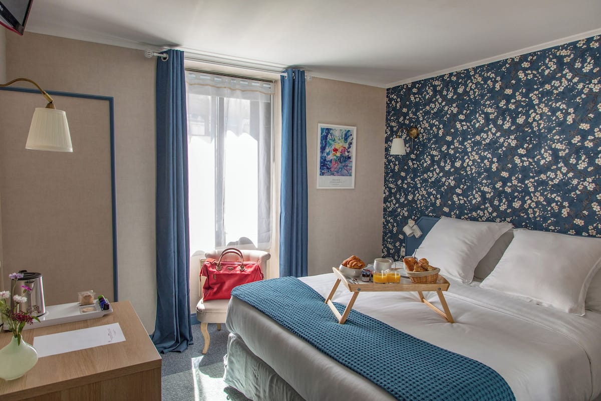 A hotel room with a bed, a desk with a lamp, a chair, a window with curtains, and a wall with a floral pattern.