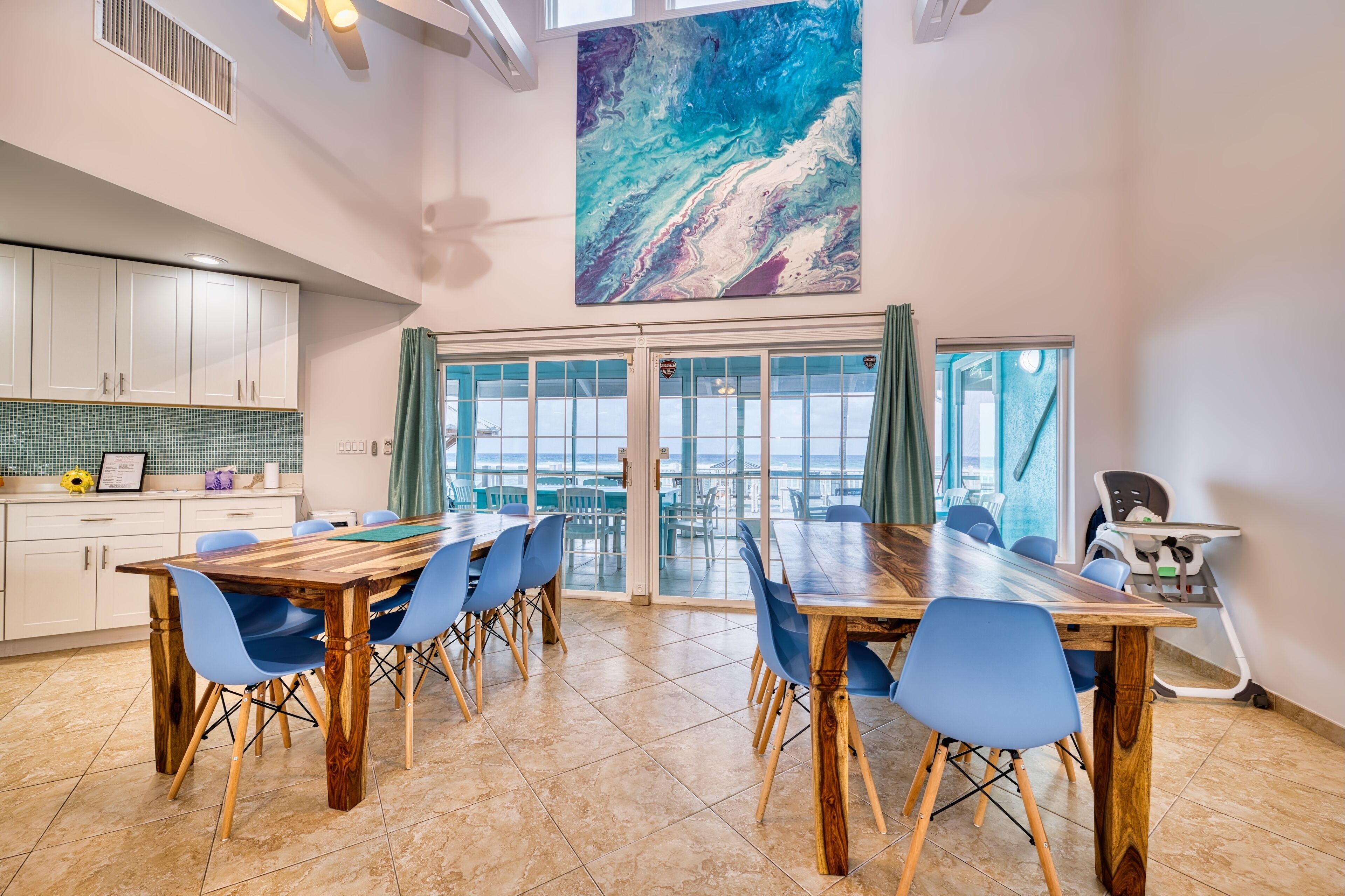 A spacious dining area with a large wooden table and blue chairs, a kitchen with white cabinets, and a large abstract painting above the windows.