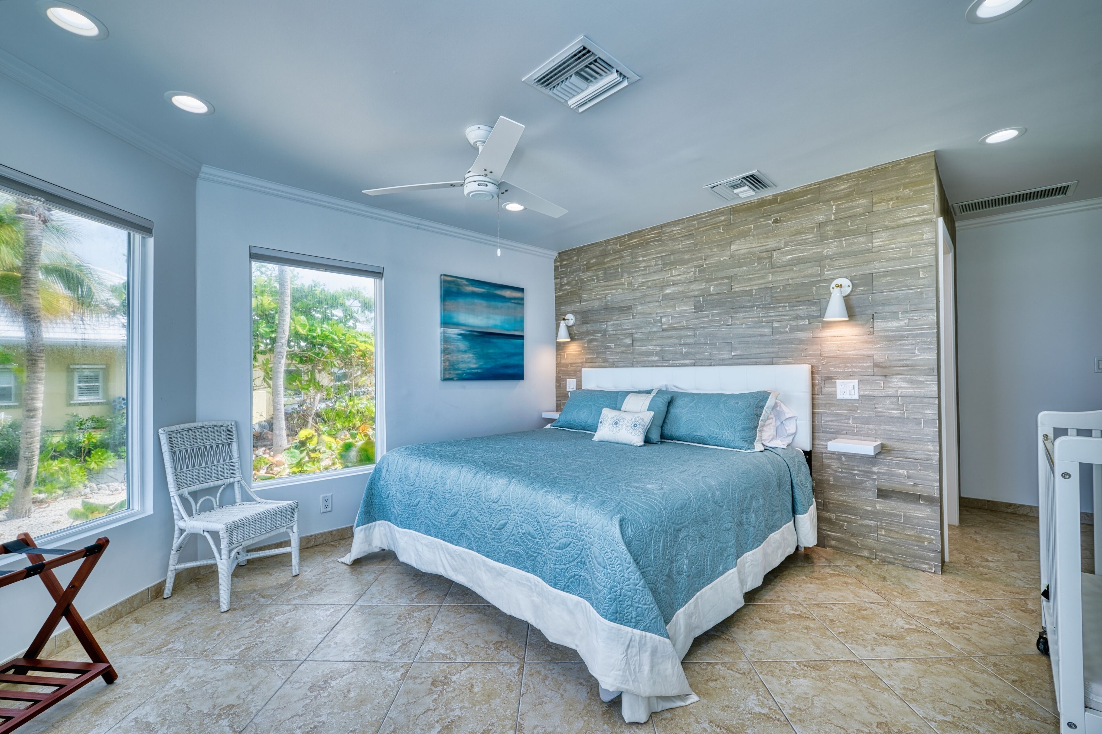 A bedroom with a large bed, a ceiling fan, a window with a view of greenery, and a brick accent wall.