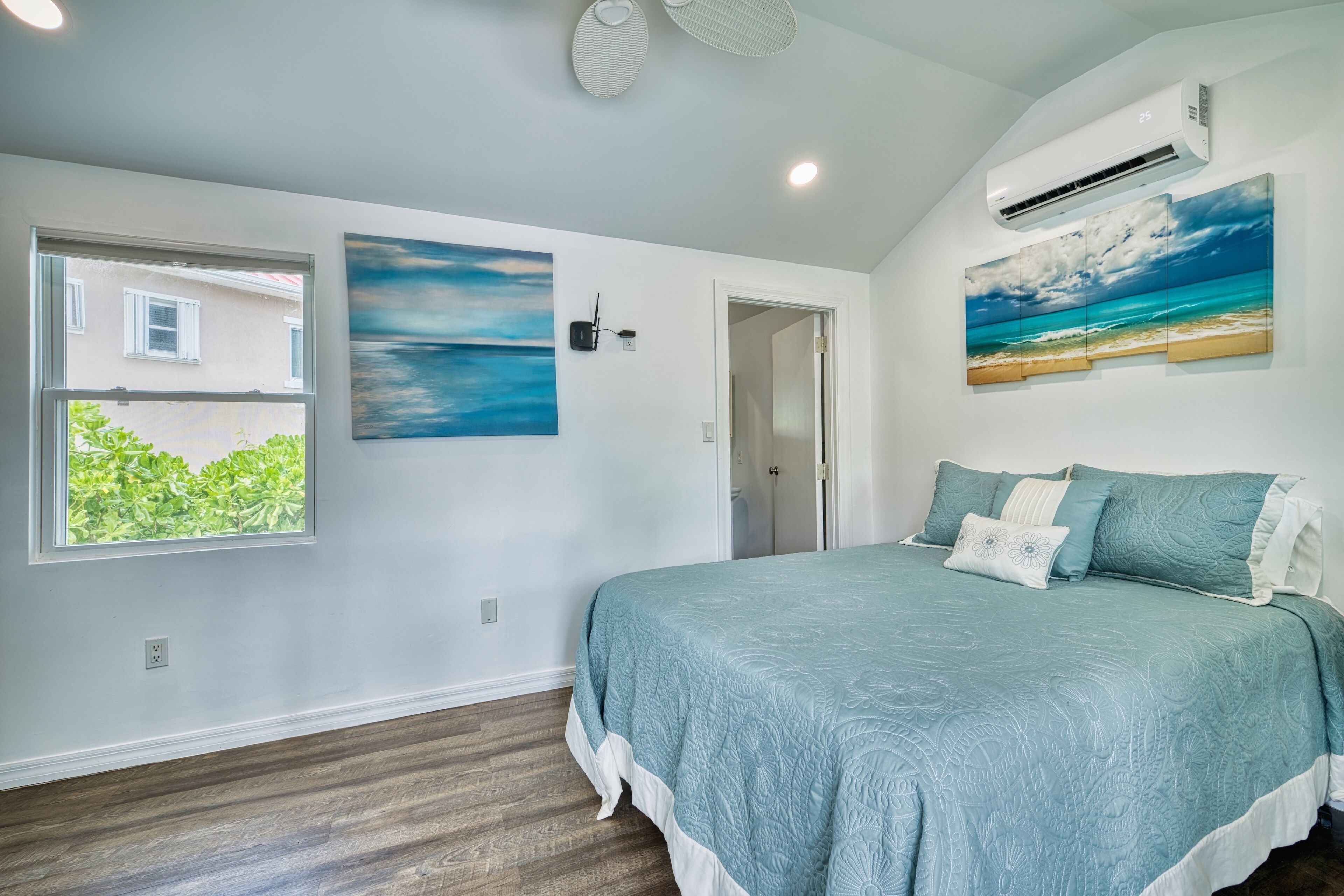 Een slaapkamer met een groot bed, een raam met uitzicht op groen en een schilderij van een strand.