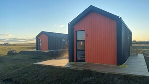 Dos cabañas modernas, rojas y negras, con terrazas de madera, ubicadas en un campo cubierto de hierba bajo un cielo despejado.