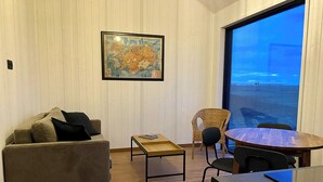 Una habitación acogedora con piso de madera, un sofá, una mesita, una silla de mimbre y una mesa de comedor con sillas. Un ventanal grande ofrece vistas al océano.