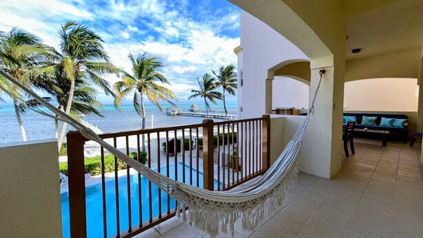 A balcony with a hammock, a pool, and a view of the ocean and palm trees. - Miramar Villas Resort (San Pedro)