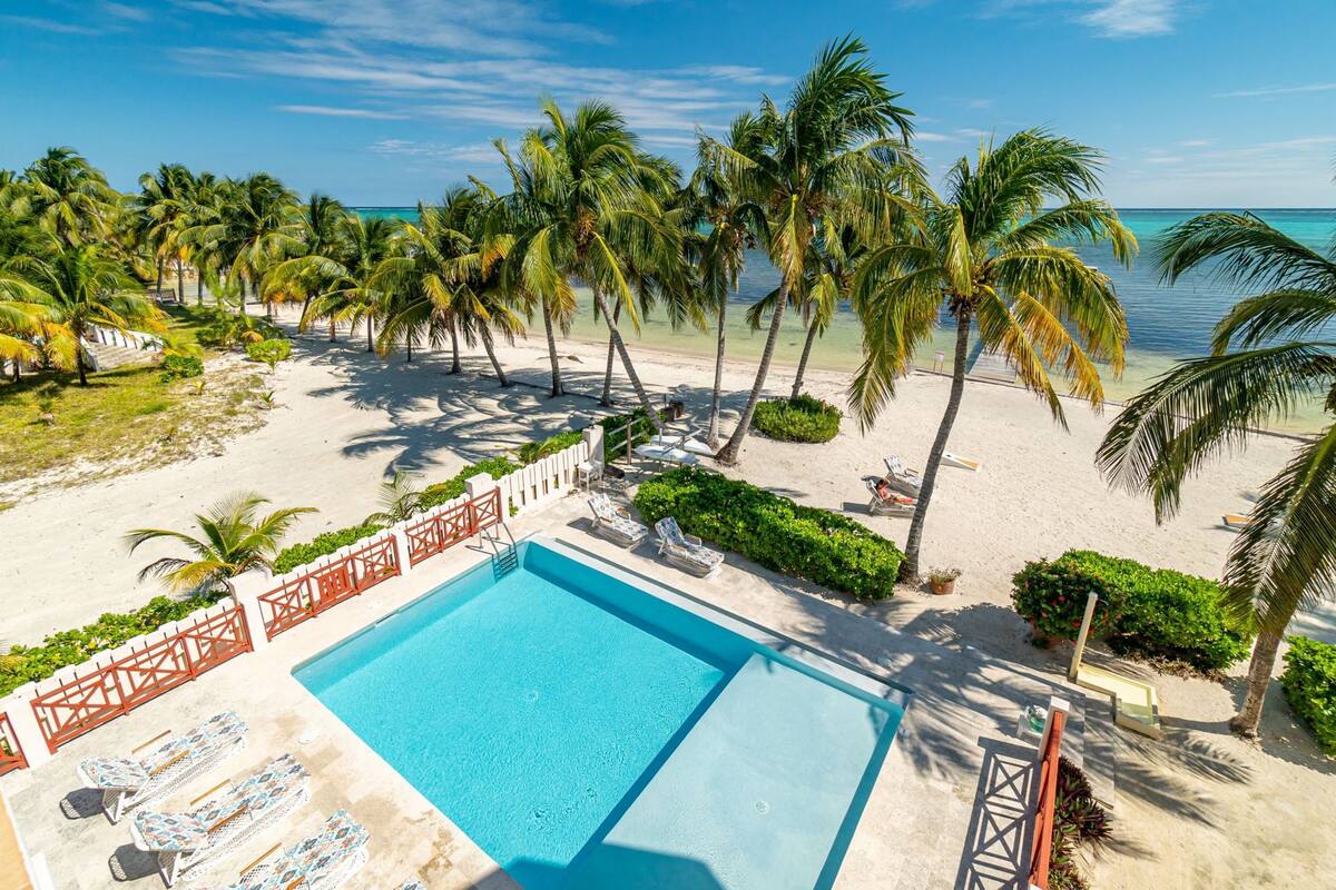 A pool area with a clear blue pool, surrounded by palm trees and a sandy beach.