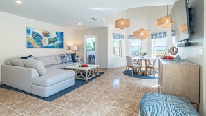 A modern living room with a sofa, coffee table, and dining area. There is a painting on the wall, a TV mounted on the wall, and a window with curtains. - Miramar Villas Resort (San Pedro)
