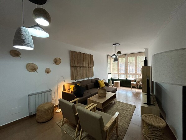 A modern living room with a sofa, armchairs, a coffee table, and a fireplace. There are hanging lights and a large window providing natural light. - Welcome to our cozy apartment in O Barco, Galicia! (Porto do Barqueiro)