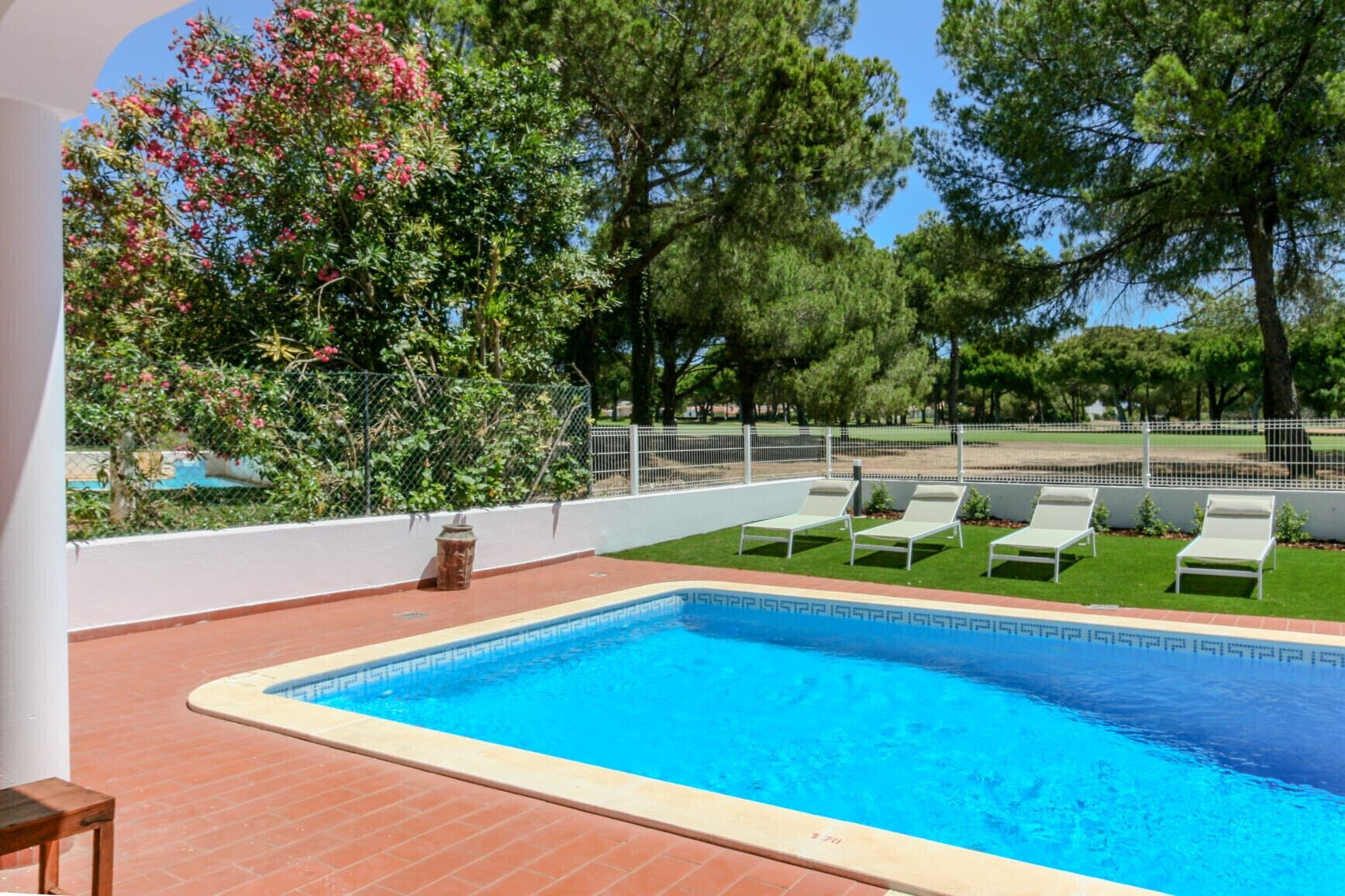 Piscina com deck revestido, cercada por uma grade branca, e várias cadeiras de descanso.