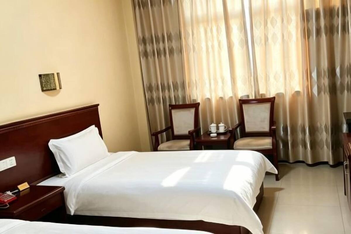 A hotel room with two beds, a nightstand, a chair, a small table, and a window with curtains.