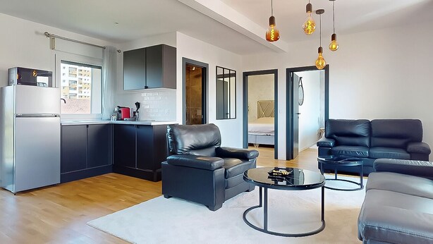 Uma sala de estar moderna com um sofá de couro preto, uma mesa de centro e uma área de cozinha com geladeira e armários.