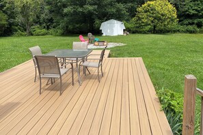 Una terraza de madera con una mesa y sillas, rodeada por un jardín cubierto de césped, un cobertizo y árboles.