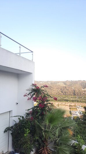 A modern building with a white facade, a balcony with metal railings, and a view of a hilly landscape in the background.