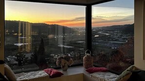 A room with a large window offering a cityscape view at sunset.