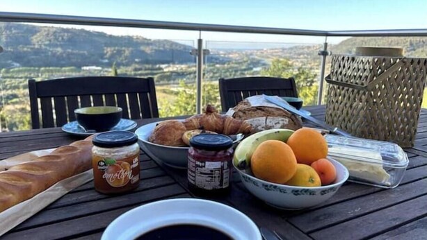 A table with breakfast items including bread, fruits, and condiments, set on a balcony with a scenic view.