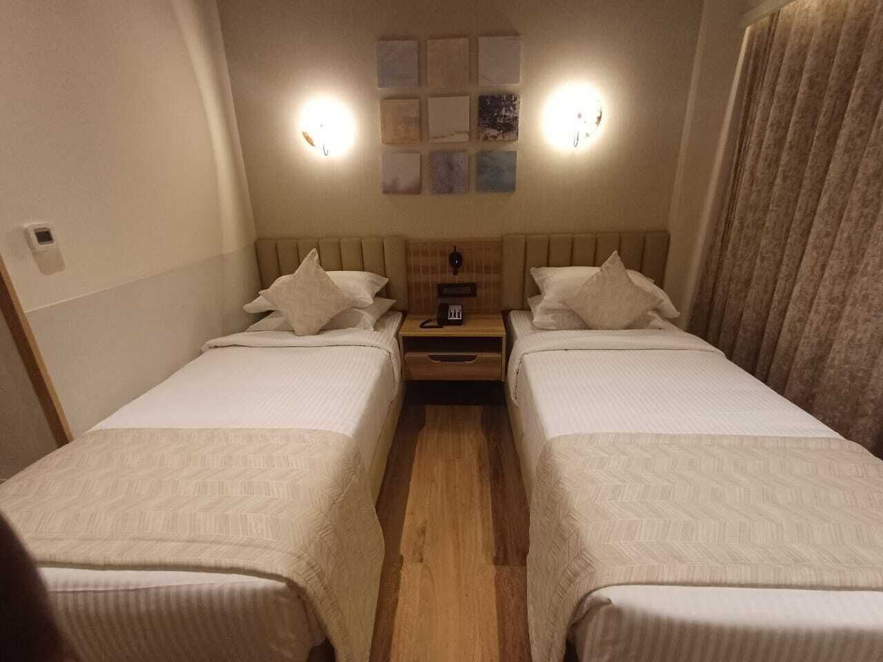 A hotel room with two beds, a wooden floor, a nightstand with a lamp, and wall-mounted artwork.