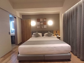 Una habitación de hotel moderna con una cama grande, una mesita de noche con lámpara y vistas al pasillo.