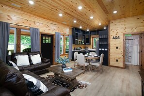 A cozy cabin interior with a living area featuring a brown leather sofa, a wooden coffee table, and a dining table with chairs. The room has wooden walls and ceiling, and there is a kitchen area visible in the background. - Helms Hollow | Hot Tub, Fire Pit, Movie Theater & Arcade (Sevierville)