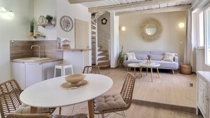 A modern kitchen with white cabinetry, a dining area with a round table, wicker chairs, and a staircase leading to a living area with a sofa and a mirror. - La Bohème 2, Mouries Centre au Cœur des Alpilles : Cosy T2 Avec Terrasse (Mouriès)