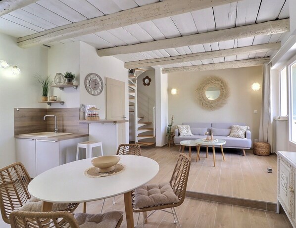 Una cocina moderna con armarios blancos, un comedor con una mesa redonda, sillas de mimbre y una escalera que lleva a una sala de estar con un sofá y un espejo.