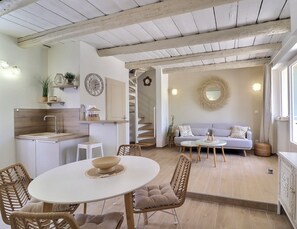 A modern kitchen with white cabinetry, a dining area with a round table, wicker chairs, and a staircase leading to a living area with a sofa and a mirror. - La Bohème 2, Mouries Centre au Cœur des Alpilles : Cosy T2 Avec Terrasse (Mouriès)