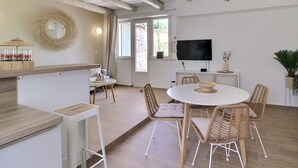 A spacious living area with a dining table, wicker chairs, a TV mounted on the wall, and a large window providing natural light. - La Bohème 2, Mouries Centre au Cœur des Alpilles : Cosy T2 Avec Terrasse (Mouriès)