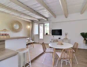 A spacious living area with a dining table, wicker chairs, a TV mounted on the wall, and a large window providing natural light. - La Bohème 2, Mouries Centre au Cœur des Alpilles : Cosy T2 Avec Terrasse (Mouriès)