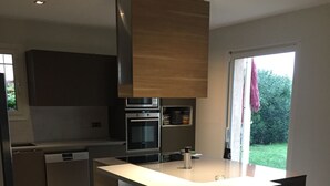 A modern kitchen with a central island, built-in oven, and a view of a garden through a sliding door. - A 15 mn de l Aèroport, Villa sur les Hauteurs de St Laurent du var (Saint-Laurent-du-Var)