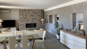 A modern living room with a stone fireplace, a flat-screen TV, a dining area with a glass table, and a kitchen area with a countertop and cabinets. - A 15 mn de l Aèroport, Villa sur les Hauteurs de St Laurent du var (Saint-Laurent-du-Var)